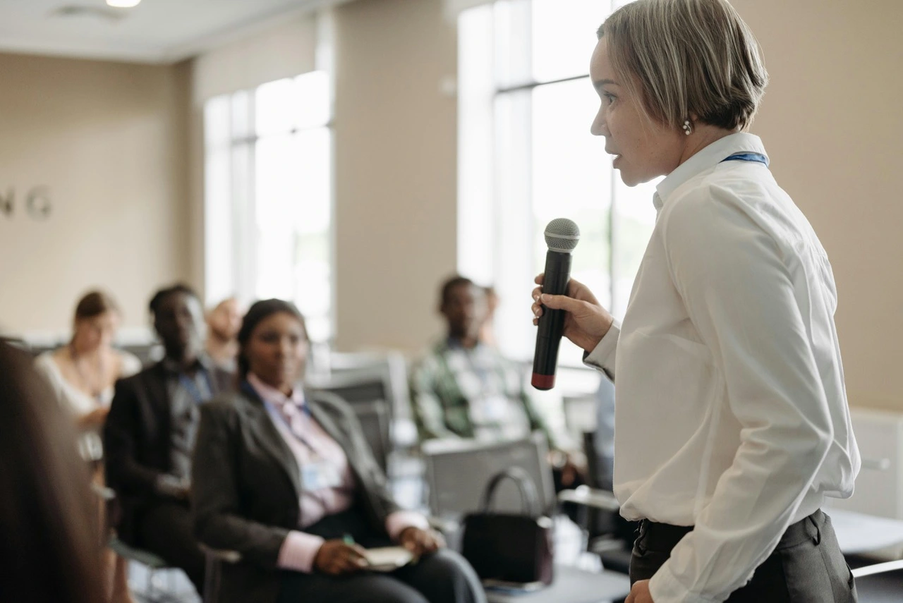 the photo of the woman with a mic speaking to a group